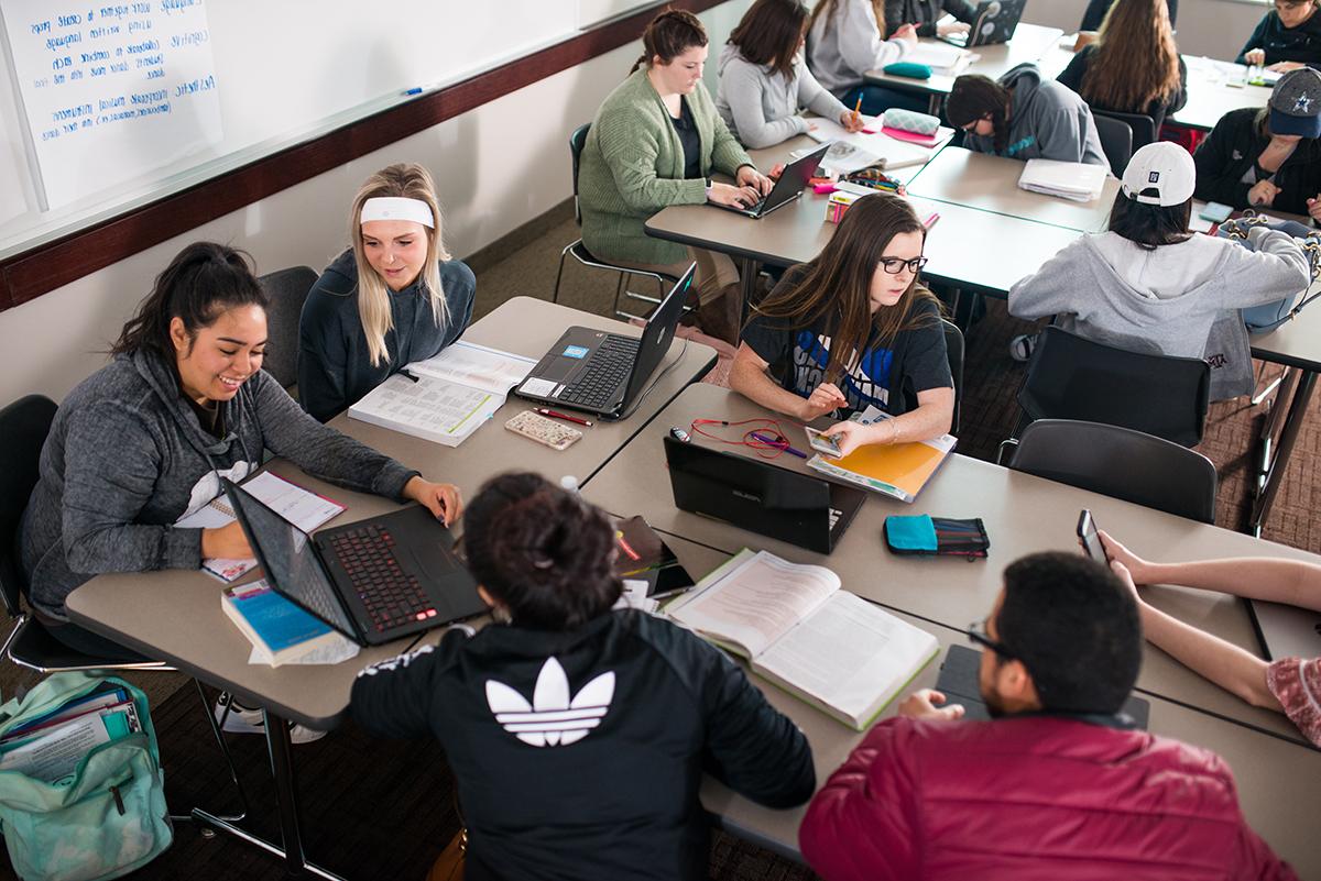 A group of students sitting down in class facing each other, using computers and talking.